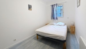 A small, bright bedroom with a single bed, a window with curtains, a small bedside table, and a radiator. - LE CLOS DE DOMINO (Saint-Georges-d'Oléron)