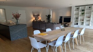 Eine moderne Küche mit Kochinsel, ein Essbereich mit Holztisch und weißen Stühlen sowie ein Wohnbereich mit Sofa und Fernseher.