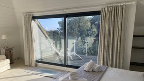 Ein Schlafzimmer mit einer großen Schiebeglaswand, einem Bett mit weißen Leinen, einem Nachttisch mit Lampe und Blick auf Gartenmöbel.