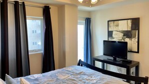 A bedroom with a bed, a TV on a desk, a ceiling fan, and a window with curtains.