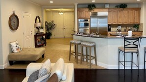 A spacious living room with a sofa, a small table, and a decorative clock. A kitchen with wooden cabinets, a stainless steel refrigerator, and a bar area is visible in the background.