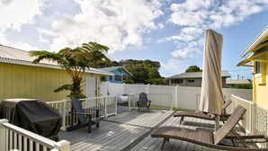 A wooden deck with lounge chairs and a grill, surrounded by a white fence and a yellow building. - Makani Ea Home 2 Bedroom Home by RedAwning (Hakalau)