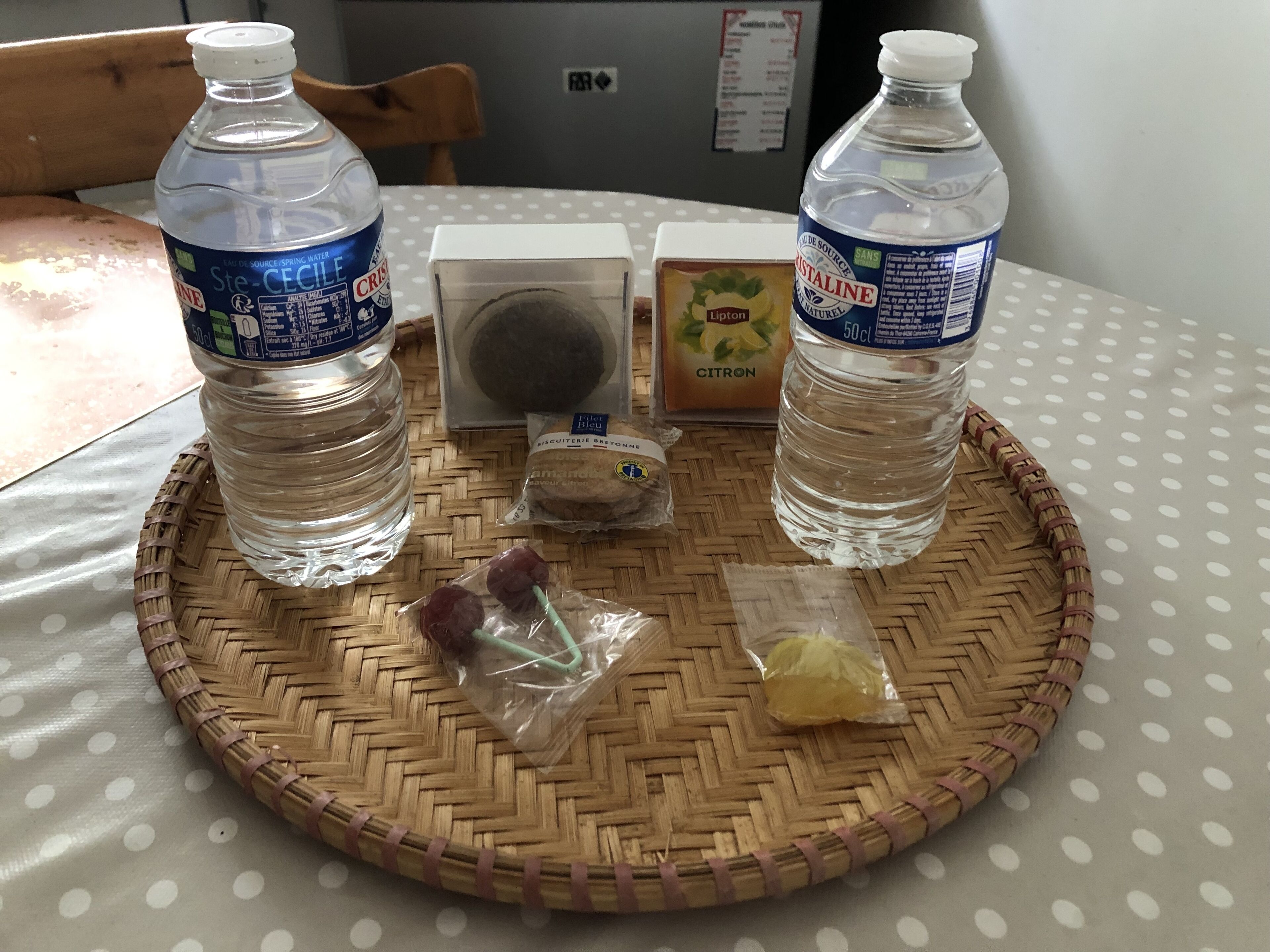 Ein Tisch mit einer gewebten Unterlage, zwei Wasserflaschen, einer Keksdose, einer Teeverpackung und einem kleinen Plastikbeutel mit Trauben.