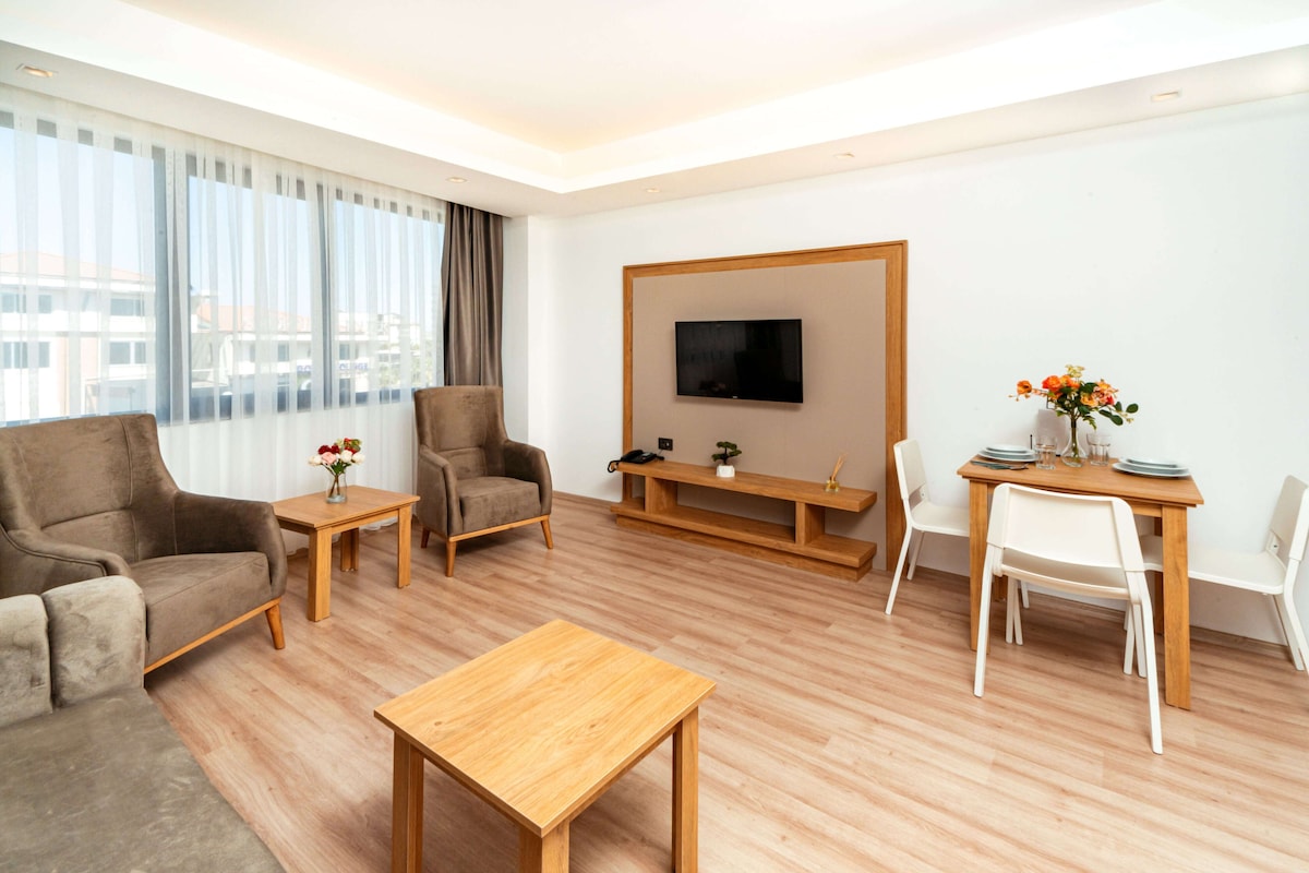 A modern hotel room with a sofa, armchair, wooden coffee table, TV, and a dining area with chairs and a small table.