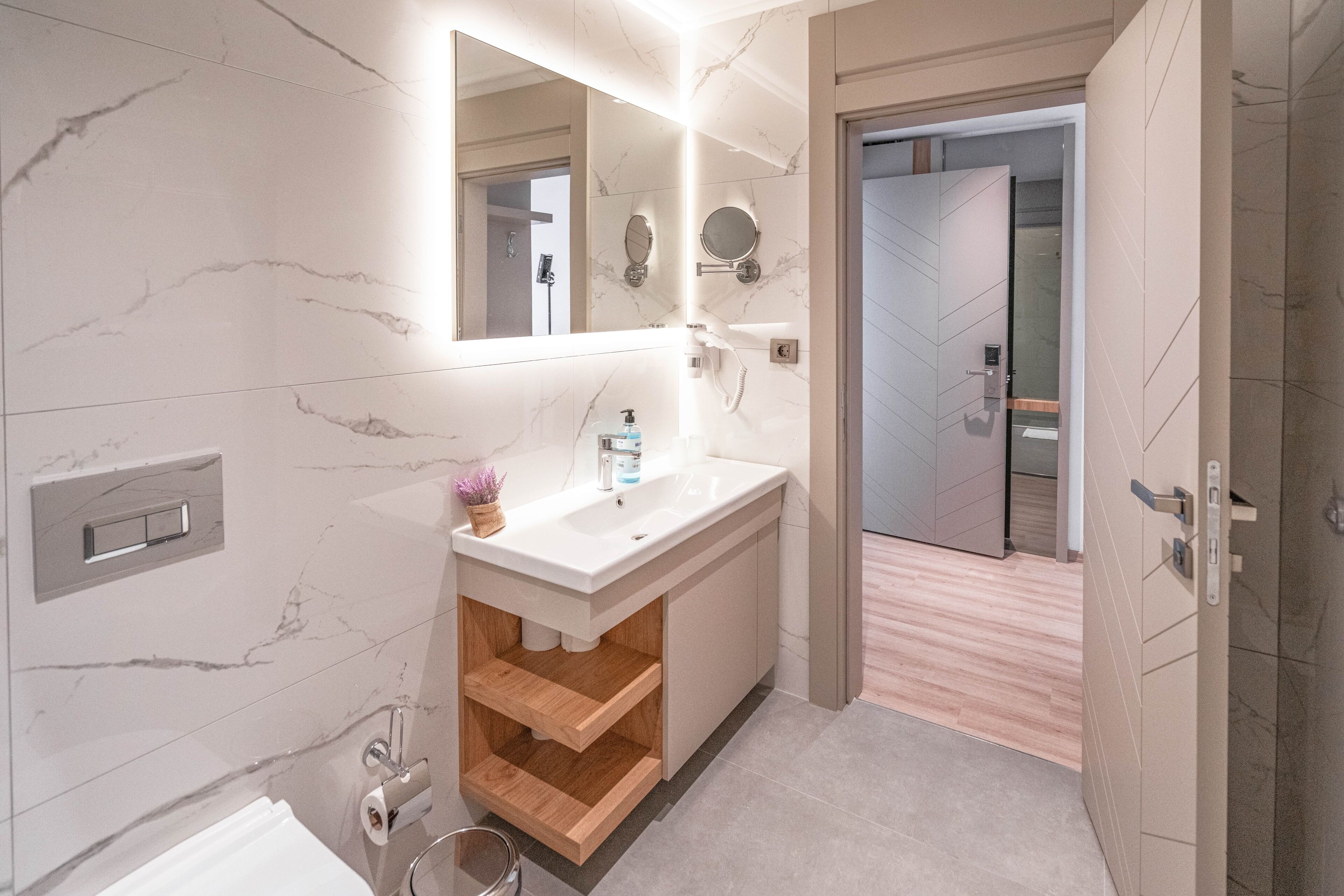 A modern bathroom with a marble vanity, a large mirror, a toilet paper holder, and a wooden shelf.