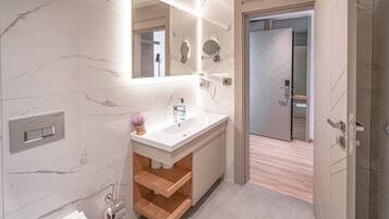 A modern bathroom with a marble vanity, a large mirror, a toilet paper holder, and a wooden shelf.
