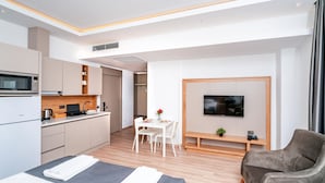 A modern hotel room with a bed, a seating area with a TV, a kitchenette, and a dining table.