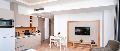 A modern hotel room with a bed, a seating area with a TV, a kitchenette, and a dining table.