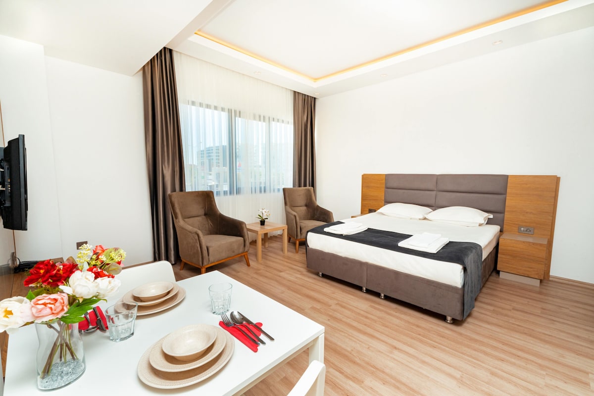 A hotel room with a bed, two armchairs, a small table with a vase of flowers, and a dining table set with plates and cutlery.