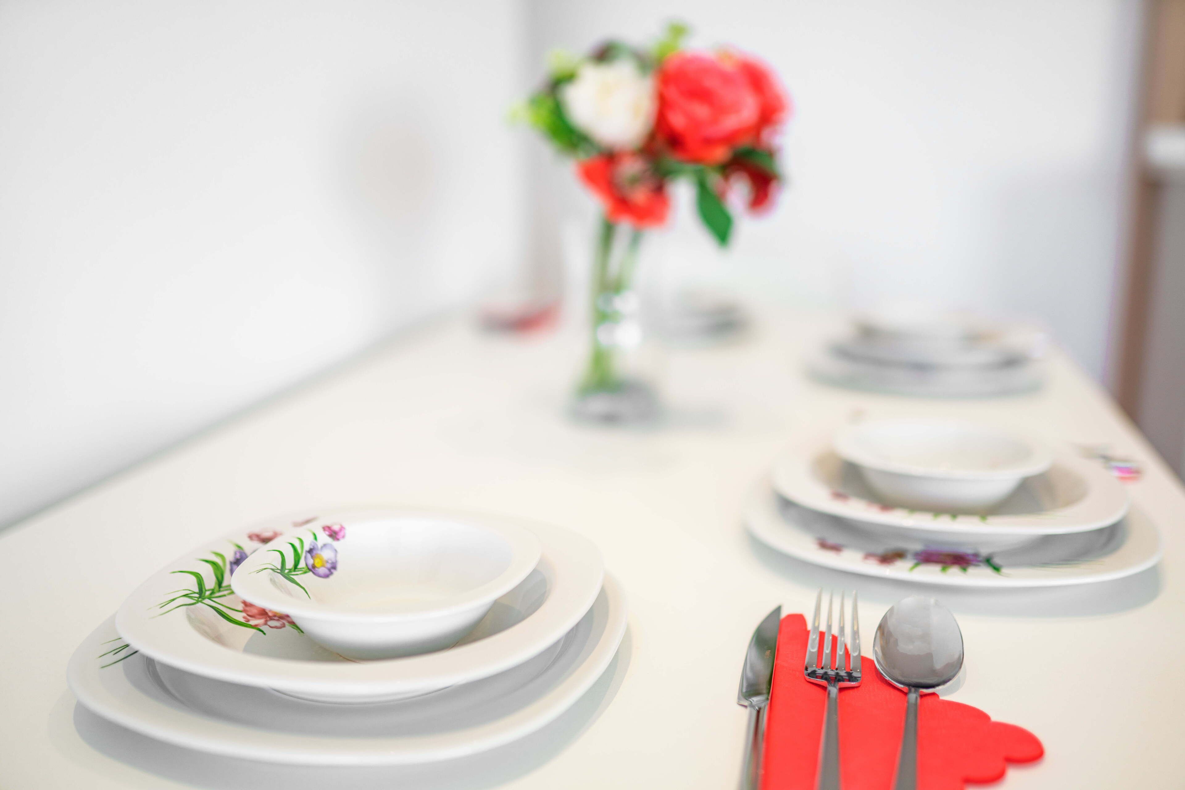 Taula de dinar ben a punt amb plats blancs, gots vermells i coberts, acompanyada d'un ram de flors.