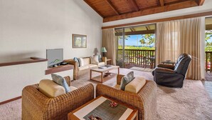 Amplia sala de estar con muebles de mimbre, un televisor de pantalla plana y un balcón con vistas a palmeras y al océano.