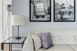 Una sala de estar moderna con un sofá, una mesa de centro de vidrio, una lámpara de pie y dos fotografías en blanco y negro enmarcadas en la pared.