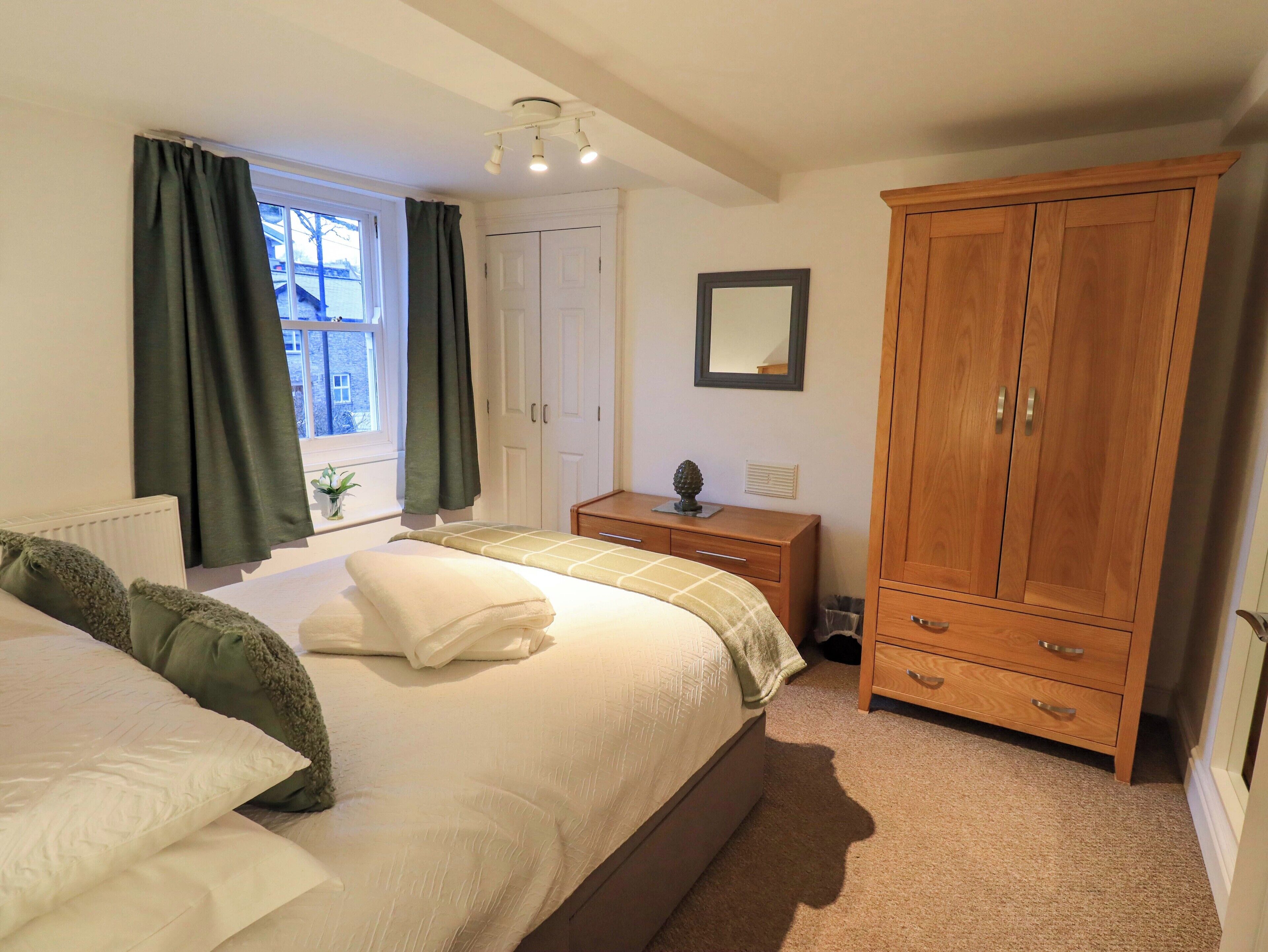 A bedroom with a large bed, a wooden wardrobe, a bedside table, and a window with curtains.