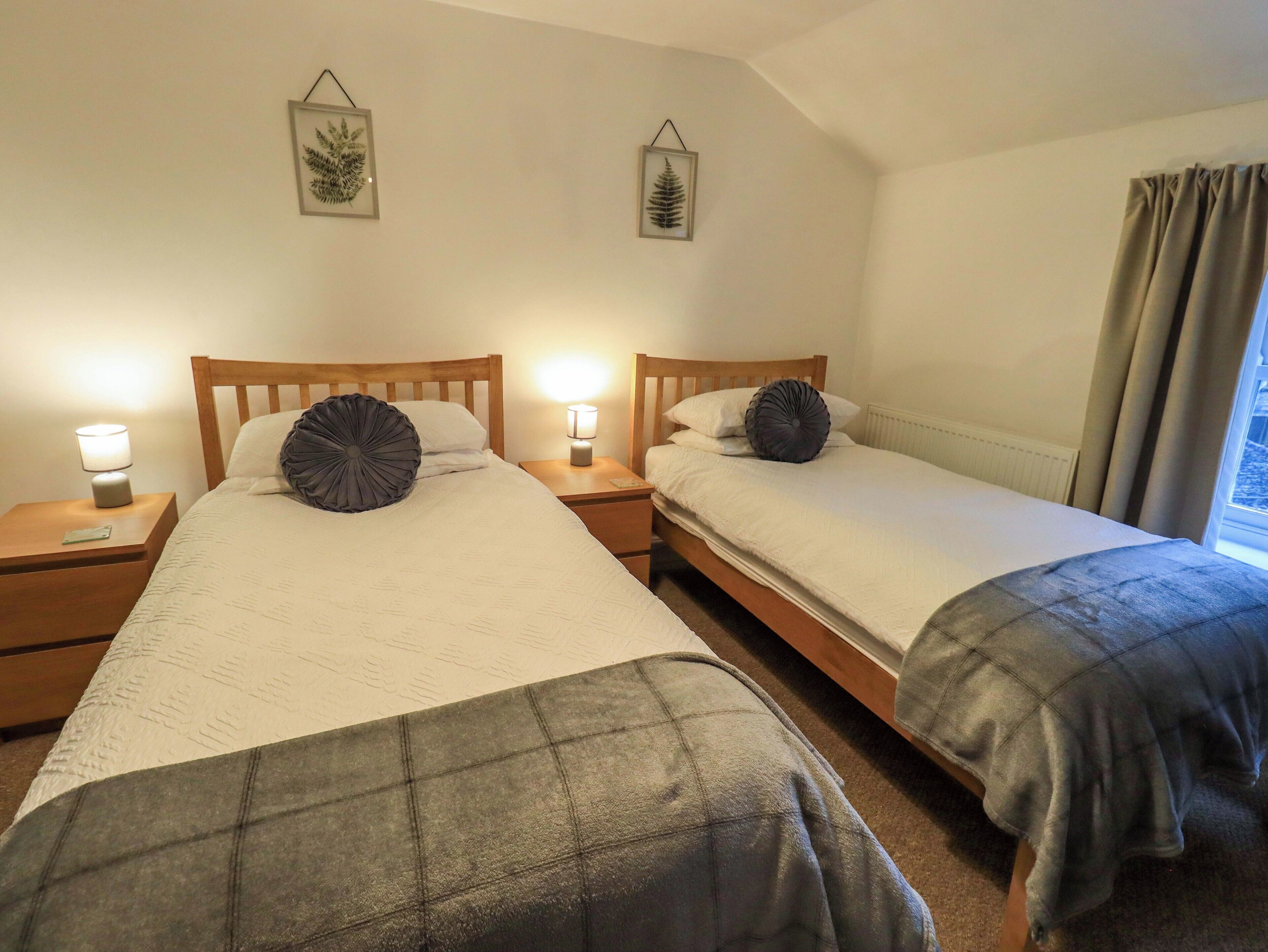A double room with two beds, each with a wooden headboard, a bedside table with a lamp, and a framed picture on the wall.
