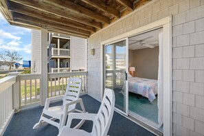 A balcony with white plastic chairs, a sliding glass door leading to a bedroom with a bed and a nightstand, and a view of a neighboring building with a balcony.