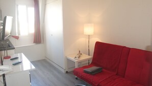 Een kamer met een rode bank, een bureau met een computer en afstandsbedieningen, een lamp en een raam met gordijnen.