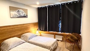 Una habitación con dos camas, cabecera de madera, radiador, una silla y una ventana con cortinas.