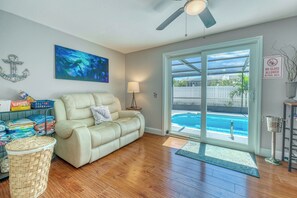 A living room with a beige sofa, a painting, a ceiling fan, and a view of a swimming pool through a glass door.
