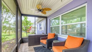 Un patio cubierto con mobiliario de mimbre y cojines naranja, ventanas amplias y un ventilador de techo.