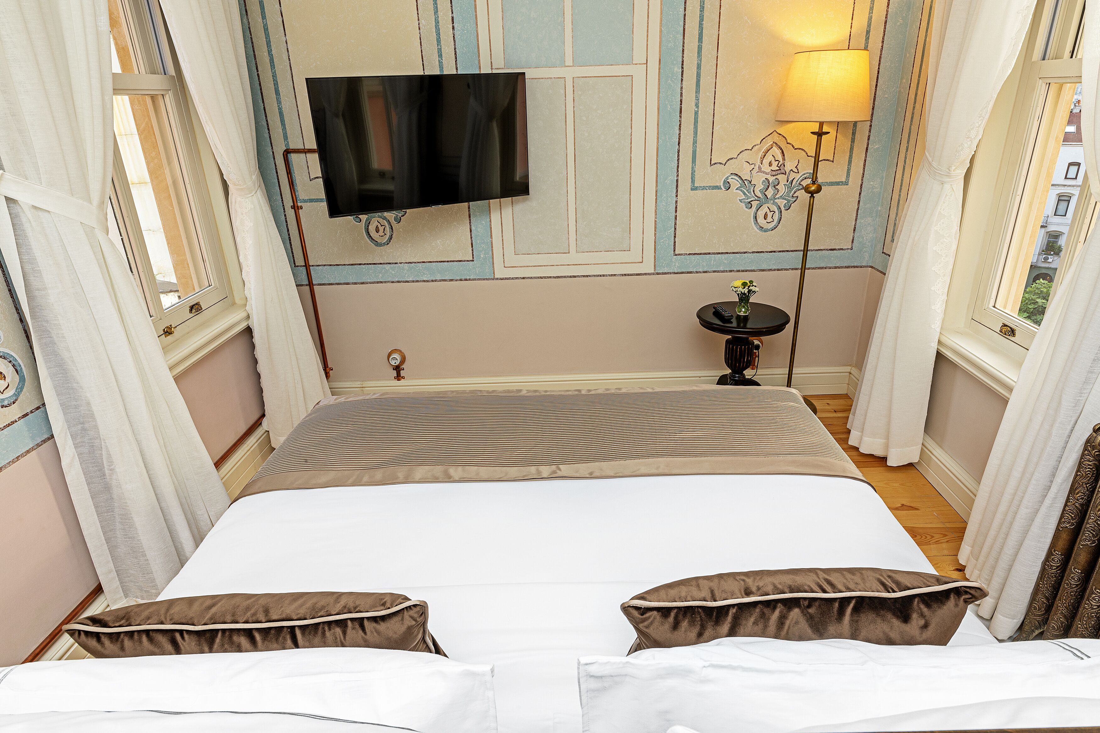 A hotel room with a large bed, a flat-screen TV mounted on the wall, a bedside table with a lamp, and a view of the city through the window.