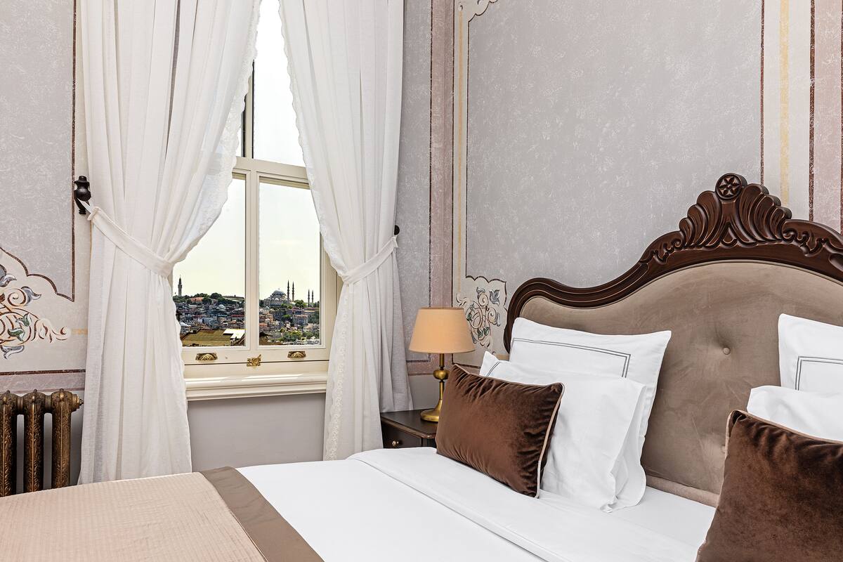 A neatly made bed with white and brown pillows, a gold lamp on a bedside table, and a view of a cityscape through a window with white curtains.