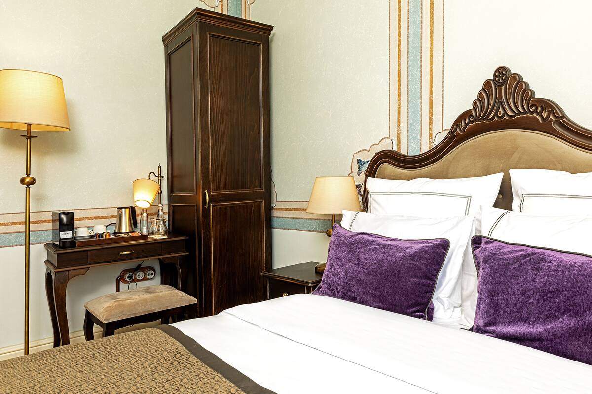 A hotel room with a large bed, two bedside lamps, a wooden wardrobe, a desk with a lamp, and a small bench.