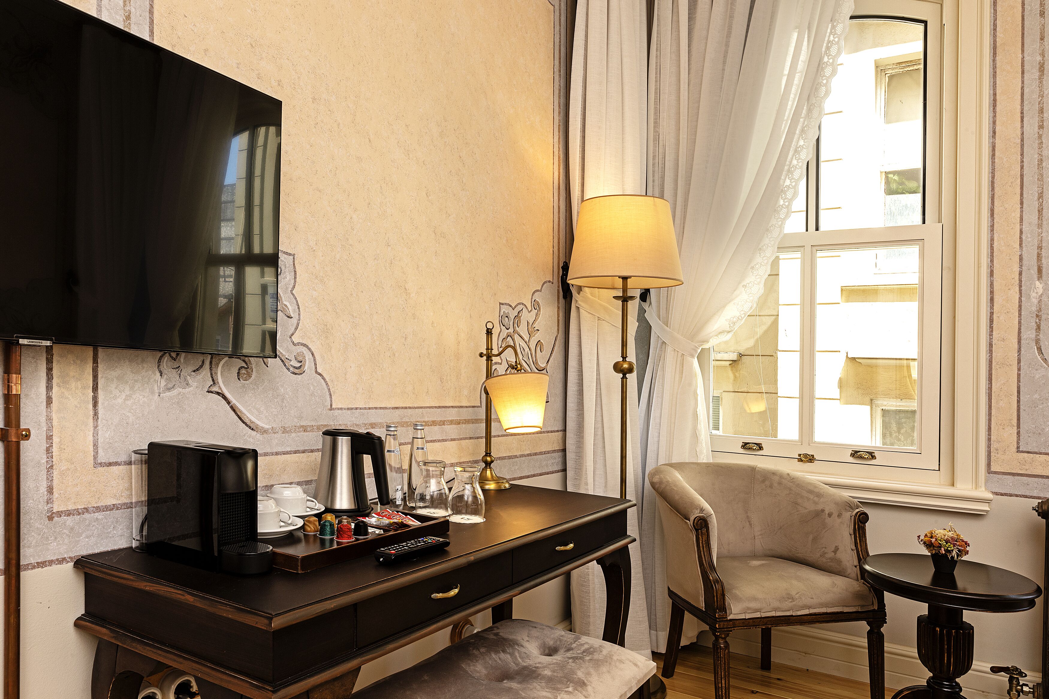 Habitación de hotel con televisión de pantalla plana, cafetera, lámpara, una silla y una mesita con un florero.