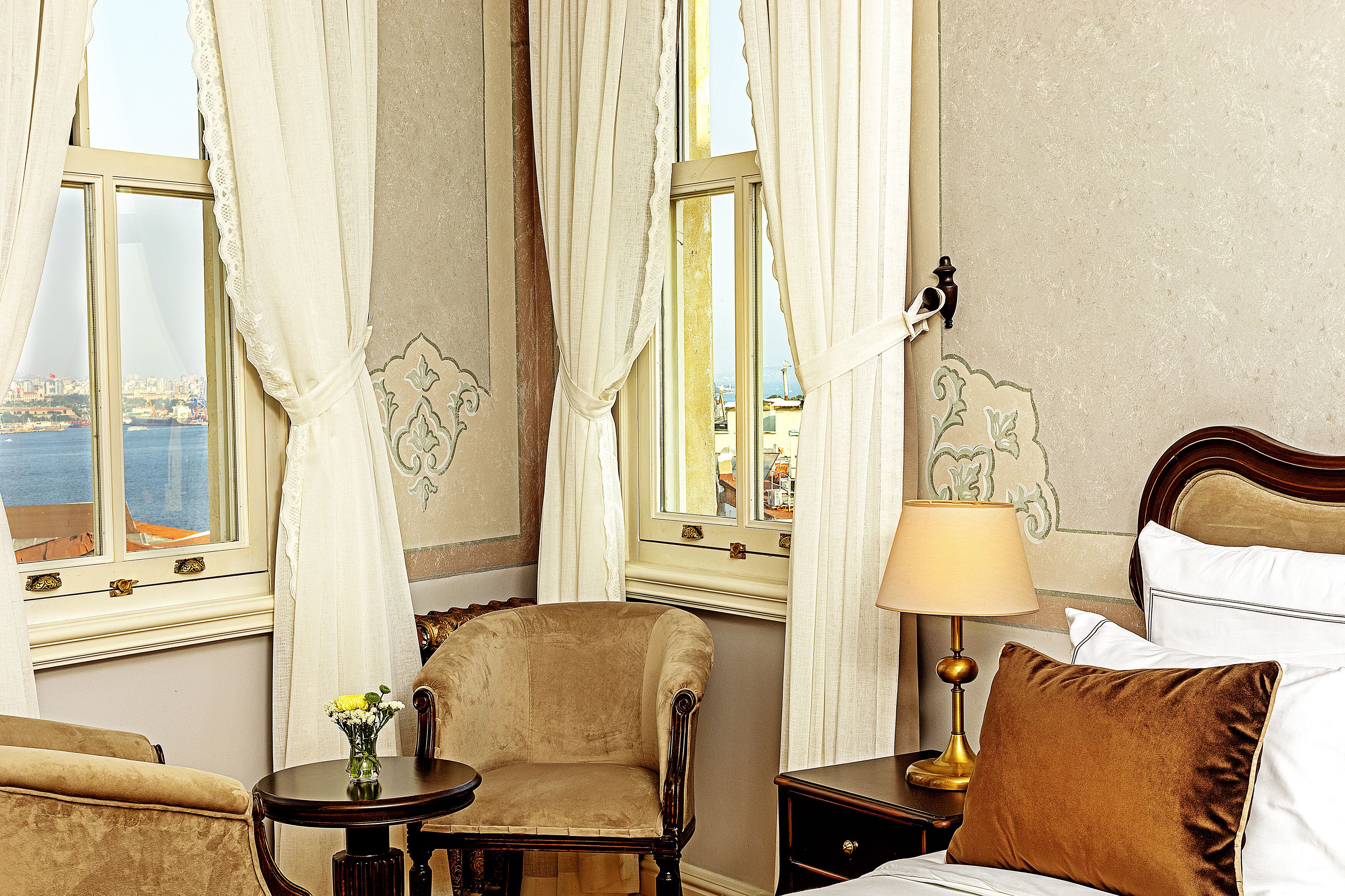 A hotel room with a view of the city, a bed with pillows, a chair, a table with a vase of flowers, and a lamp.