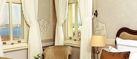 A hotel room with a view of the city, a bed with pillows, a chair, a table with a vase of flowers, and a lamp.