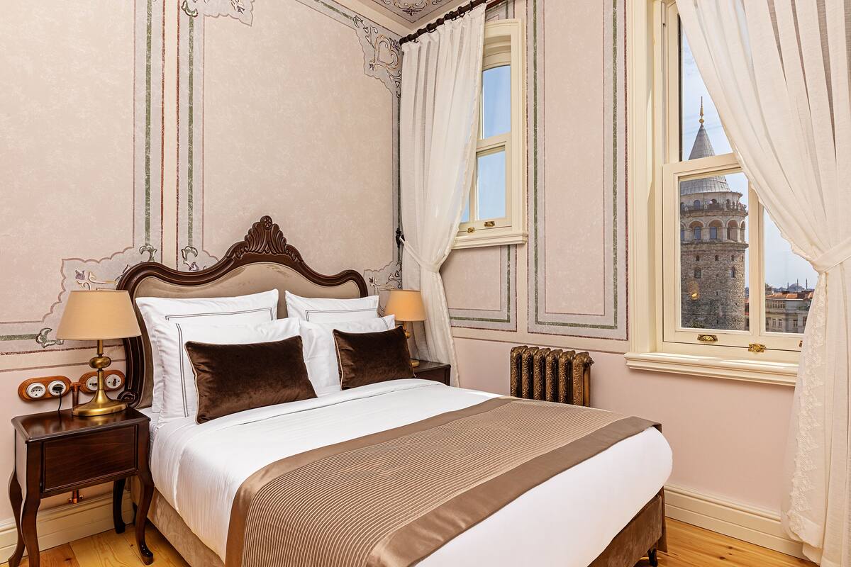 A neatly made bed with white and brown linens, a wooden headboard, and two bedside lamps. A window with curtains reveals a view of a historic building.