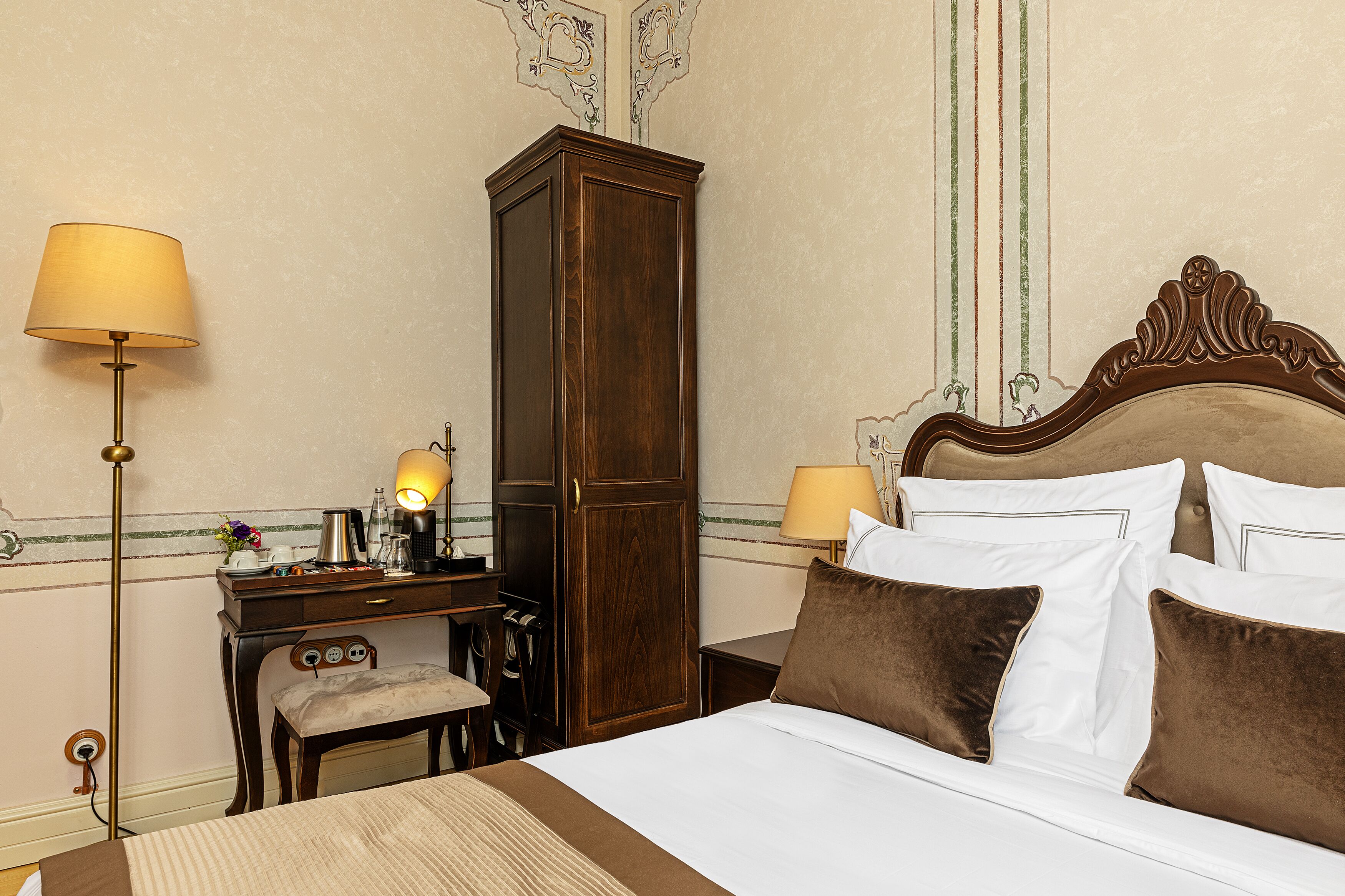 A hotel room with a bed, bedside lamps, a wooden wardrobe, a desk with a lamp, and a chair.