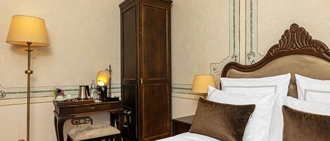 A hotel room with a bed, bedside lamps, a wooden wardrobe, a desk with a lamp, and a chair.
