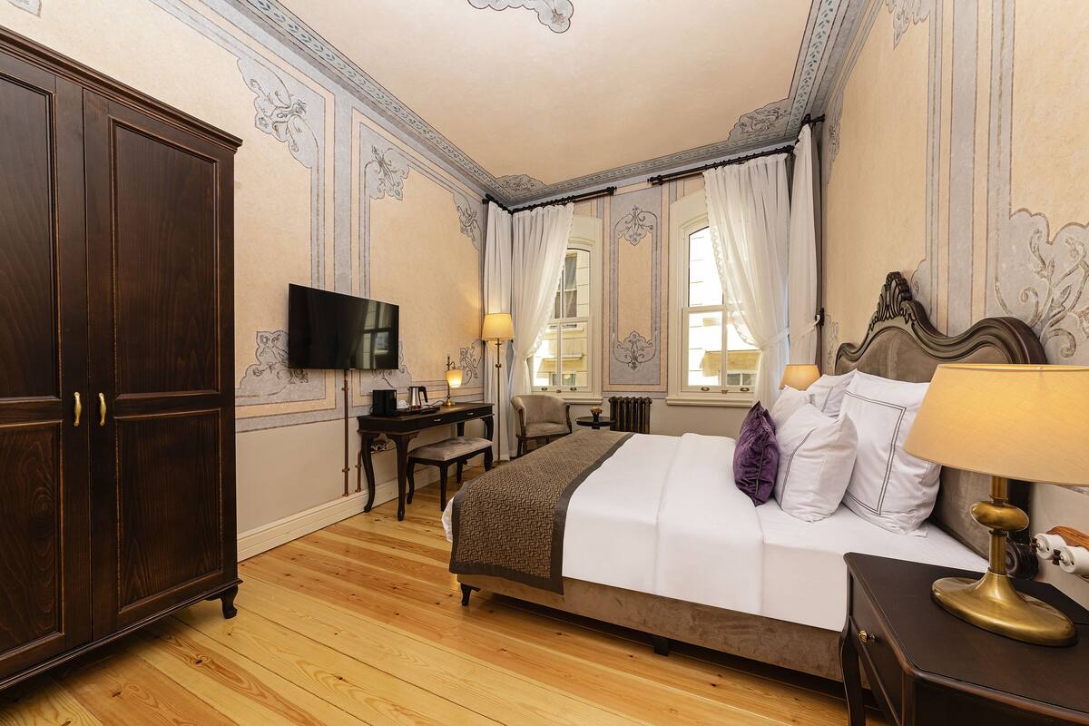 A hotel room with a large wooden wardrobe, a desk with a chair, a bed with white bedding and a decorative headboard, and a television mounted on the wall.