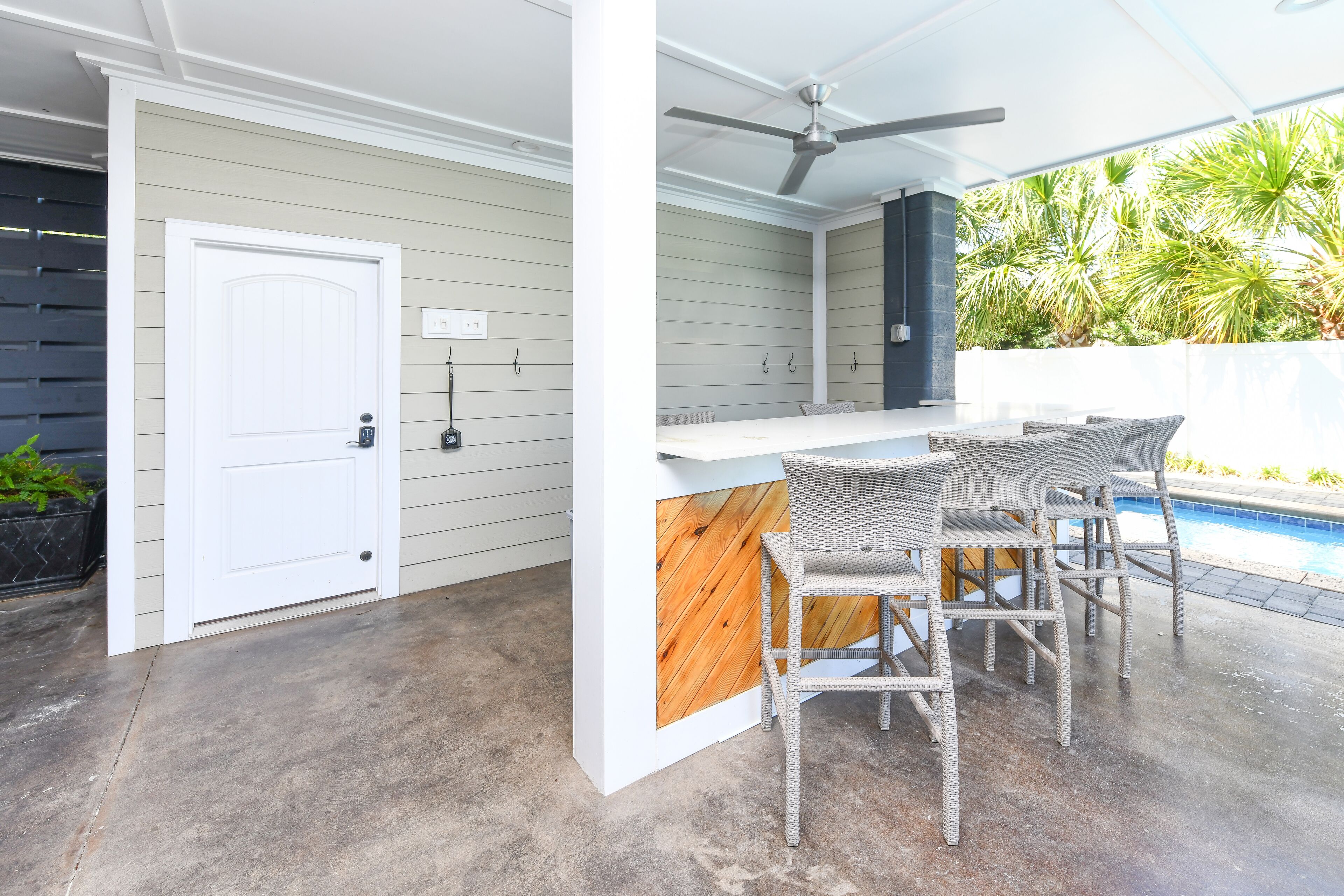 A covered outdoor area with a bar counter, two wicker chairs, a ceiling fan, and a pool in the background.