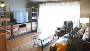 A living room with a television, wooden furniture, a glass coffee table, and a large window with curtains.
