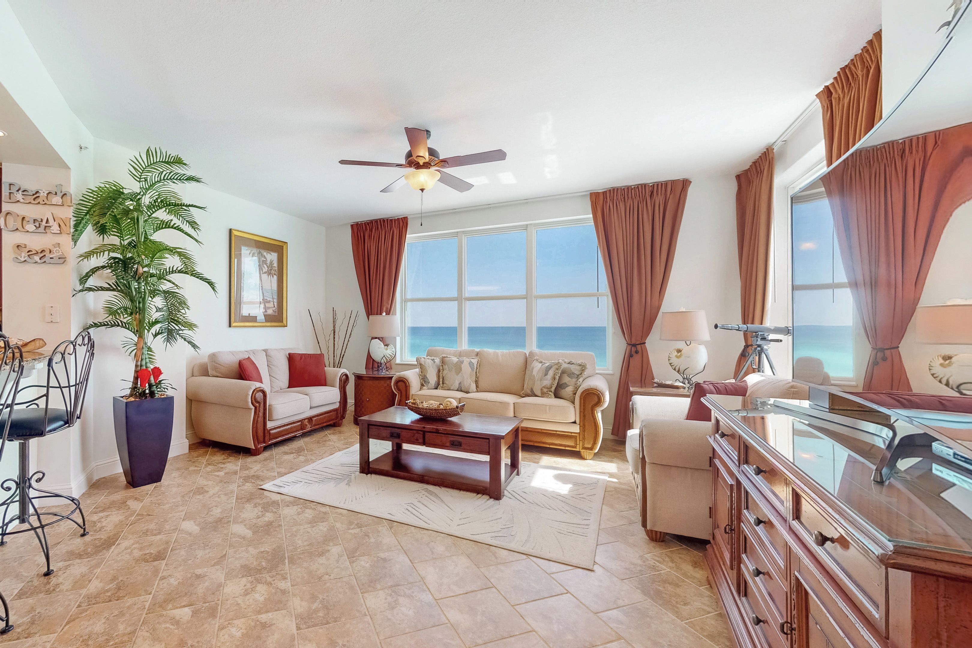 A spacious living room with a ceiling fan, large windows with curtains, a coffee table, and a view of the ocean.