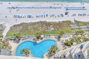 Vue aérienne d’un complexe hôtelier en bord de mer, avec une grande piscine entourée de palmiers et de chaises longues.