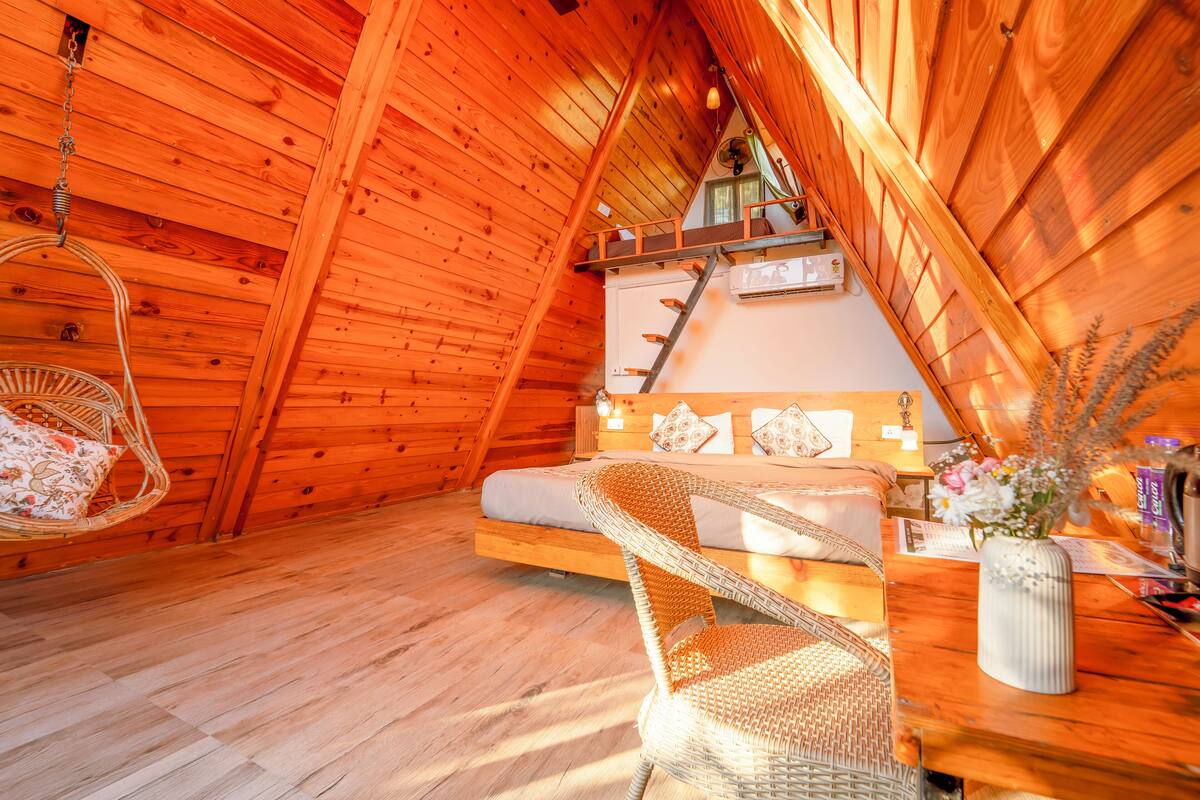 A cozy wooden cabin with a loft bedroom, a hanging chair, and a wooden dining table with a vase of flowers.