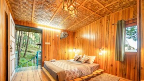 A wooden cabin bedroom with a bed, nightstands, and a view of the forest. - Paradise Pool Resort (Nainital)