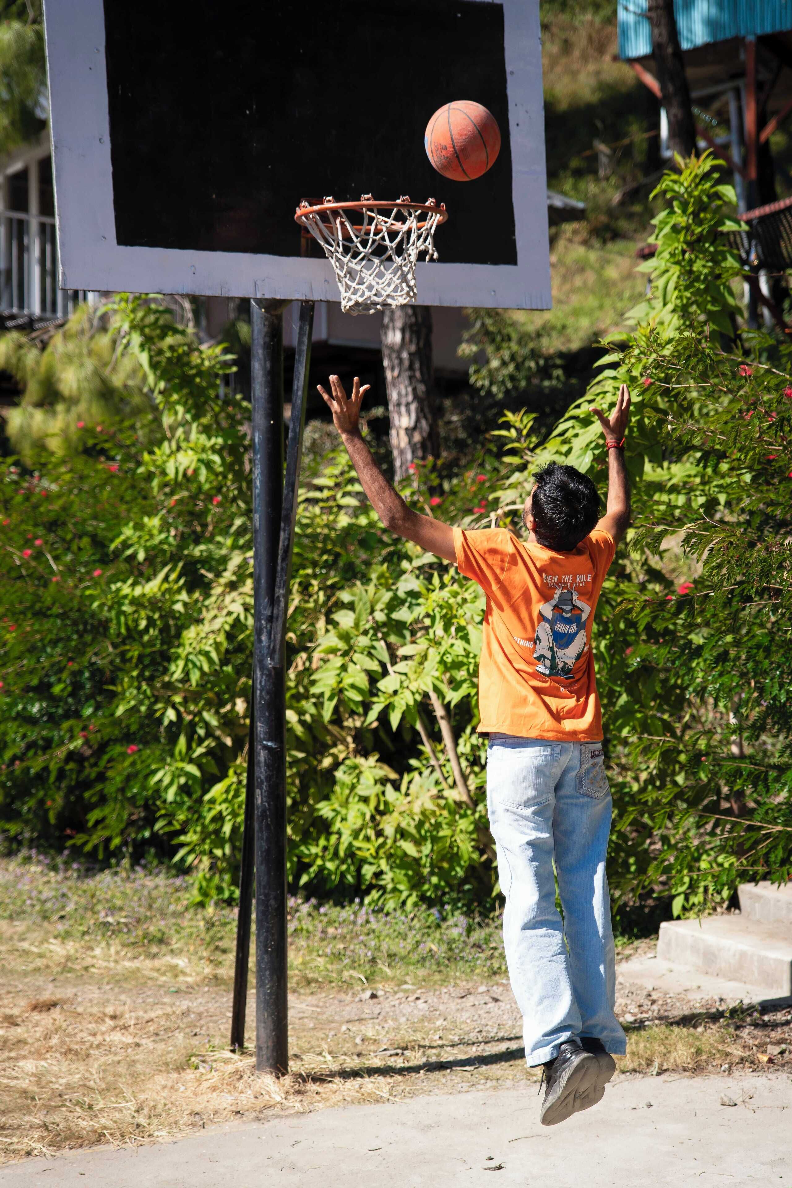 Un aro de baloncesto con red, un balón de baloncesto en pleno vuelo, y una persona con camiseta naranja y vaqueros alcanzando la pelota.