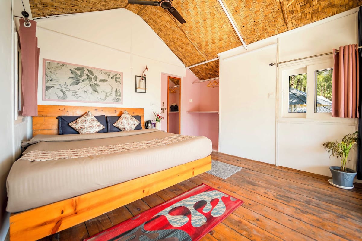 A bedroom with a wooden bed frame, a large bed, a wooden ceiling, a window with curtains, and a potted plant.