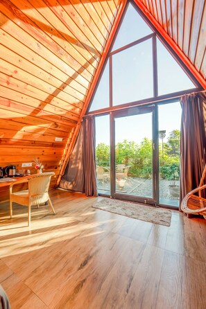 A cozy, wooden-paneled room with a large window, a dining table, and a wicker chair. - Paradise Pool Resort (Nainital)