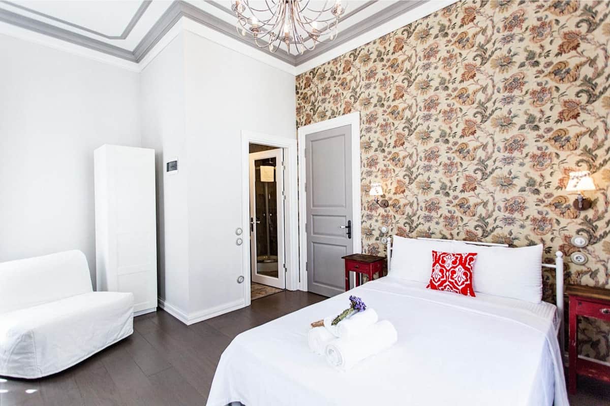 Habitación de hotel con una cama grande, un sillón blanco, zona de ducha y pared con estampado floral.