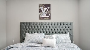 A bedroom with a tufted headboard, patterned bedding, and a framed artwork on the wall.