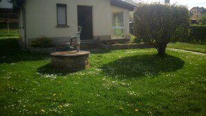 A single-story house with a stone well in the front yard, surrounded by a well-maintained lawn and a small tree. - Quiet vacation home with garden (Lichtenstein/Sa.)