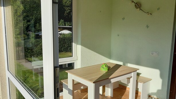 A room with a wooden table and bench, a window with a view of greenery, and a wall-mounted light fixture. - Quiet vacation home with garden (Lichtenstein/Sa.)