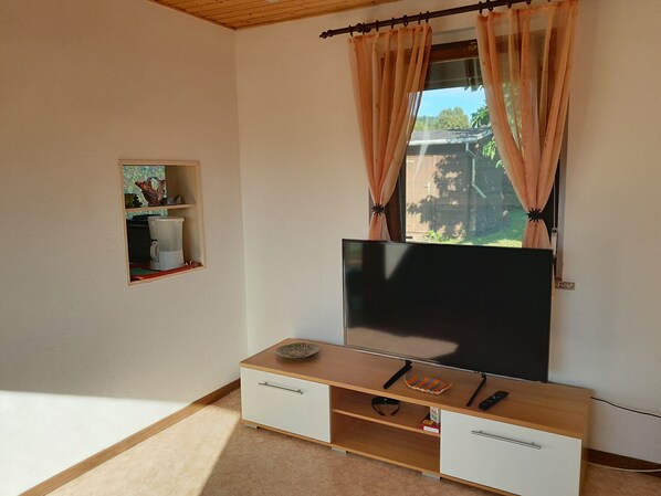 A room with a TV on a wooden stand, a small shelf with decorative items, and a window with curtains. - Quiet vacation home with a garden (Lichtenstein/Sa.)