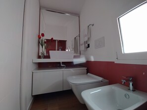 A bathroom with a white sink, a white toilet, and a bidet. There is a mirror above the sink and a window on the right side.
