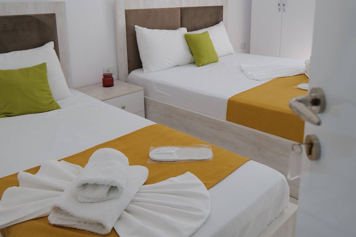 A hotel room with two beds, each with a wooden headboard, white bedding, and a mustard-colored bed runner. There is a nightstand with a red lamp between the beds, and a white door is visible in the foreground.