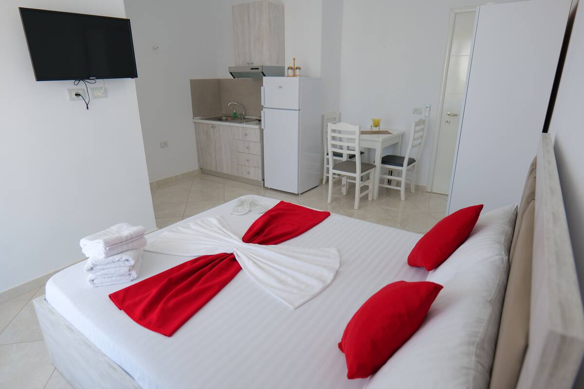 A hotel room with a bed, a TV mounted on the wall, a kitchenette, and a dining area.